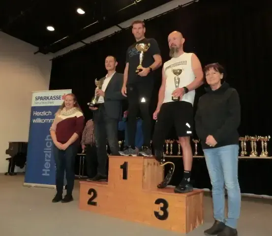 Eine Gruppe von Menschen steht auf einem Podest mit Nummern und hält Trophäen. Sie lächeln und posieren für ein Foto. Das Podest hat drei Ebenen, mit der Nummer 1 oben. Dahinter ist eine Wand mit einem blauen Banner.