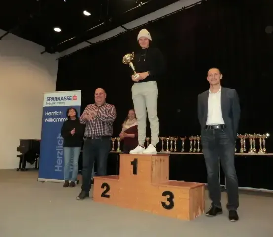 Eine Frau steht mit einer Trophäe auf einem Podest, umgeben von anderen, einschließlich zwei Männern. Sie befinden sich in einem Raum mit einem blauen Banner, Trophäen und einem Klavier.