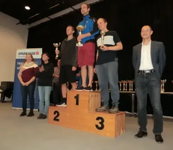 Fünf Personen stehen auf einem Podium mit den Nummern 1, 2 und 3. Sie halten Pokale. Dahinter steht ein Banner mit dem Wort 'Sparkasse'. Ein Klavier und mehrere Pokale sind im Hintergrund.