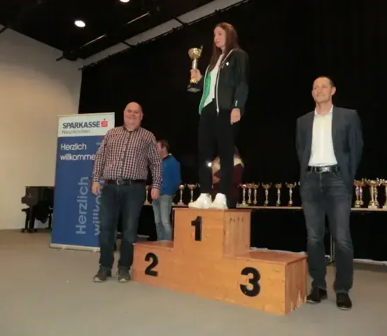 Eine Frau steht auf einem Podium und hält eine Trophäe. Zwei Männer stehen neben ihr auf niedrigeren Podesten. Ein Banner mit den Worten 'Sparkasse' und 'Herzlich willkommen' ist im Hintergrund sichtbar.