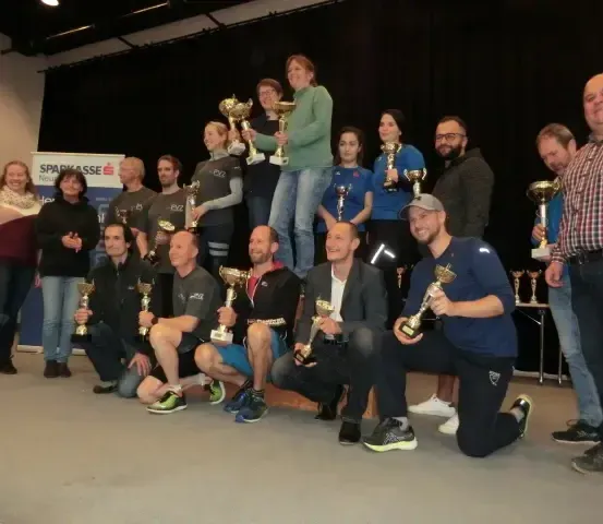 Eine Gruppe von Menschen posiert mit Trophäen für ein Foto, eine Frau in Grün steht auf den Schultern einer anderen Person.