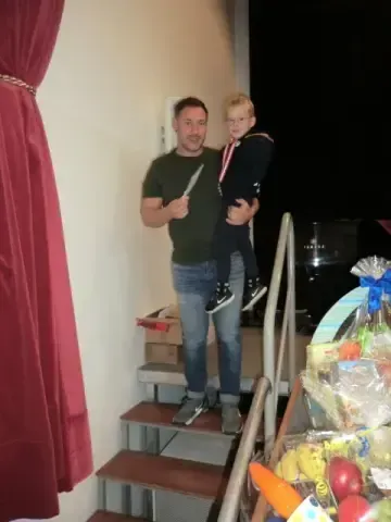 Ein Mann hält ein Messer in der Hand, während er mit einem Kind auf seinen Schultern auf einer Treppe steht. Rechts steht ein Korb mit Obst. Dahinter befindet sich eine Wand mit einem Vorhang und Kisten.