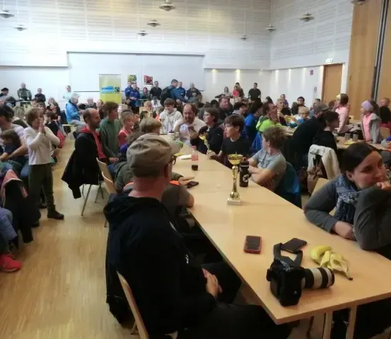 Ein großer Raum mit Tischen und Stühlen ist mit vielen Menschen gefüllt, einige sitzen und andere stehen. Ein Mann mit Hut sitzt an einem Tisch mit einem Pokal, und eine Frau sitzt in der Nähe mit Bananen und einer Kamera.