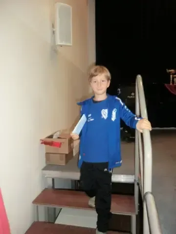 Ein junger Junge in einem blauen Shirt steht auf einer Treppe, hält ein weißes Papier in der Hand und lächelt. Hinter ihm stehen Kisten auf einem Tisch, und eine Lautsprecher ist an der Wand montiert.