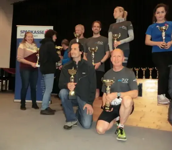 Eine Gruppe von Menschen, darunter Männer und Frauen, posiert für ein Foto, während sie Pokale in der Hand halten. Sie stehen und knien auf einer Plattform. Sie tragen alle Turnschuhe. Hinter ihnen befindet sich ein Banner mit der Aufschrift 'Sparkasse'. Es gibt eine Holzkonstruktion mit einem Banner darauf. Im Hintergrund befindet sich ein schwarzer Vorhang.