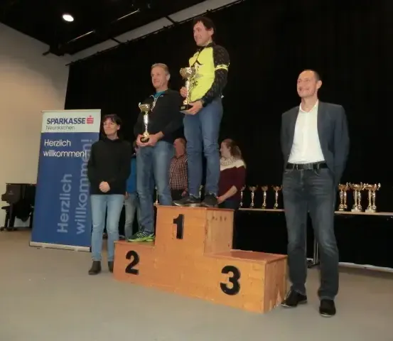 Ein Mann steht mit einer Trophäe auf einem Podium, zusammen mit zwei anderen, einer hält eine Trophäe. Hinter ihnen steht ein Banner mit der Aufschrift 'Herzlich willkommen'. Hinter dem Banner steht ein Klavier.