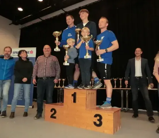 Mehrere Personen stehen mit Trophäen auf einem Podest, lächeln und posieren für ein Foto. Sie tragen blaue Hemden, Jeans und Sneakers. Dahinter steht ein Tisch mit weiteren Trophäen.