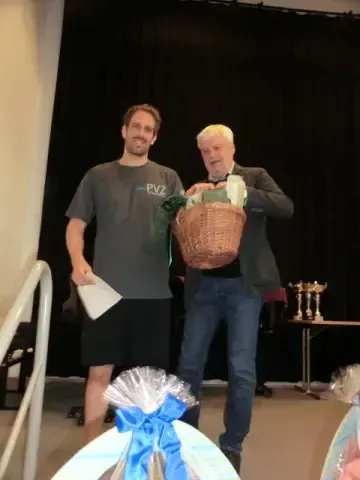 Zwei Männer stehen vor einer Bühne mit schwarzem Hintergrund, einer hält einen Korb, der andere ein Papier. Sie lächeln beide.