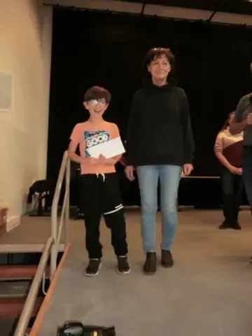 Eine Frau und ein Junge stehen lächelnd auf einer Bühne und halten ein Zertifikat. Hinter ihnen steht eine weitere Person in der Nähe eines schwarzen Vorhangs.