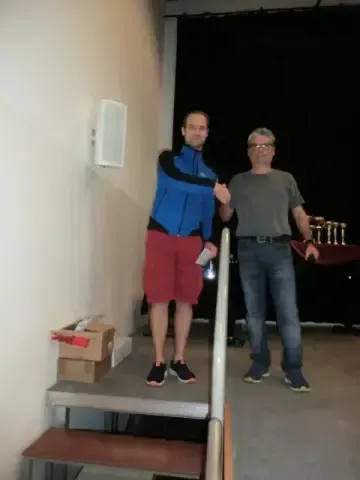 Zwei Männer stehen auf einer Bühne, einer hält eine Trophäe und präsentiert wahrscheinlich eine Auszeichnung. Die Umgebung umfasst eine Wand, einen Lautsprecher, Kisten und einen Tisch mit Bechern.