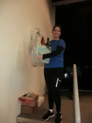 Eine Frau steht auf einer Treppe, hält eine Geschenktüte und eine Flasche und trägt ein blaues Shirt und schwarze Hose. Kisten stehen auf der Treppe, und ein weißes Geländer befindet sich rechts.