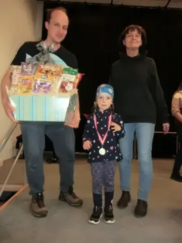 Ein Mann, eine Frau und ein junges Mädchen stehen lächelnd auf der Bühne. Der Mann hält einen Geschenkkorb mit verschiedenen Snacks. Das junge Mädchen trägt eine Medaille und Turnschuhe.
