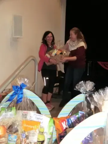 Zwei Frauen, eine mit einem Korb voller Lebensmittel und die andere mit einem Blumenstrauß, stehen vor einer Treppe mit verpackten Geschenken und verschiedenen Snacks.