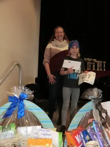 Ein junges Mädchen hält ein Stück Papier und einen Zettel in der Hand und steht neben einer Frau, beide lächeln. Sie stehen vor einem dekorierten Tisch mit verschiedenen Geschenktüten.