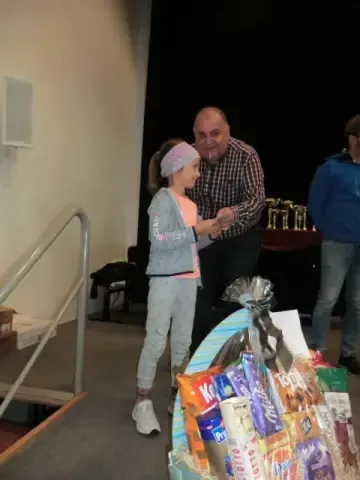 Ein junges Mädchen gibt einem Erwachsenen die Hand. Hinter ihnen steht ein Tisch mit Trophäen. Im Vordergrund befindet sich ein Korb mit verschiedenen Snacks.