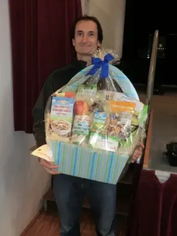 Ein Mann hält einen bunten Geschenkkorb voller Lebensmittelprodukte. Er lächelt und scheint sich bei einer Veranstaltung zu befinden. Hinter ihm sind ein roter Vorhang und eine Treppe zu sehen.