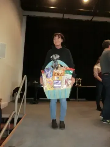 Eine Frau steht auf einer Bühne und hält einen Korb mit Snacks und anderen Gegenständen, mit Leuten im Hintergrund.