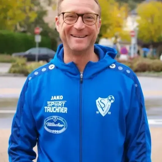 Ein lächelnder Mann in einem blauen Jackett mit verschiedenen Logos steht auf einer Straße, umgeben von Bäumen und einem verschwommenen Hintergrund.