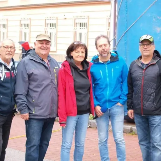 Fünf Personen in Winterkleidung stehen vor einem Gebäude. Der Mann auf der linken Seite trägt eine Brille, während die Frau in der Mitte die Hände in den Taschen hat. Der Mann auf der rechten Seite trägt eine Kappe. Dahinter befindet sich eine blaue Wand.