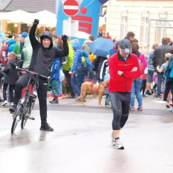 Ein Mann in Schwarz feiert mit seinem Fahrrad bei einer überfüllten Outdoor-Veranstaltung. Ein anderer Mann in Rot steht neben ihm mit verschränkten Armen. Ein Hund läuft hinter ihnen.