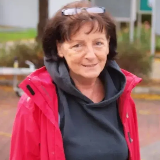 Eine Frau mit kurzen Haaren, die eine rote Jacke und ein schwarzes Shirt trägt, lächelt in die Kamera. Sie hat eine Brille auf dem Kopf. Hinter ihr ist ein verschwommener Hintergrund von einer Metallstruktur, Pflanzen und einem Rasenbereich.