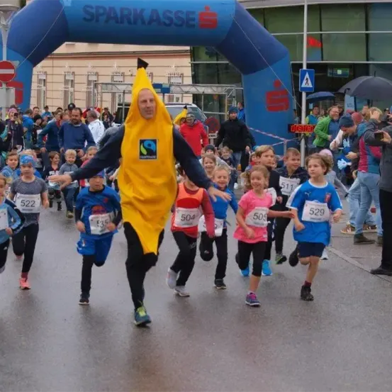 Ein Mann in einer Bananen-Kostüm führt eine Gruppe von Kindern, die an einem Marathon teilnehmen. Sie tragen nummerierte Startnummern und laufen in der Nähe eines blauen Bogens mit dem Wort 'SPARKASSE' darauf. Zuschauer beobachten von den Seitenlinien.