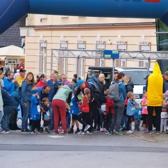 Eine Gruppe von Menschen steht in einer Schlange vor einem Gebäude mit einem blauen Bogen, während andere dahinter zusehen. Einige tragen Rucksäcke und nummerierte Trikots. Ein Auto ist in der Nähe geparkt.