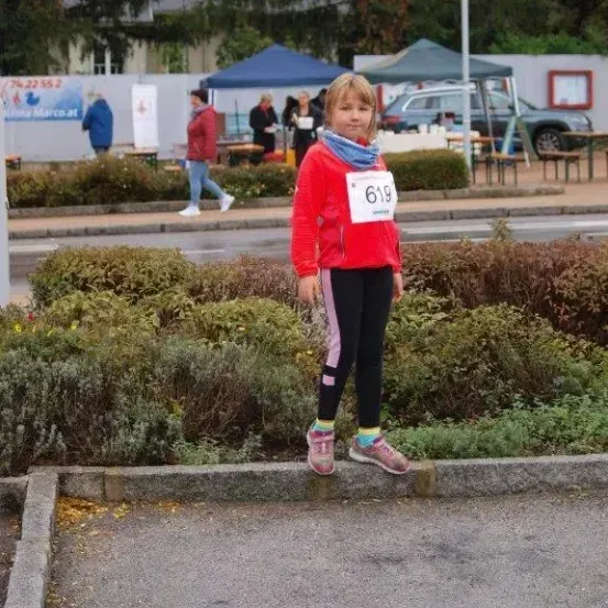 Ein junges Mädchen in einer roten Jacke steht auf der Straße, mit der Nummer 619 auf ihrem Shirt. Hinter ihr gehen mehrere Personen unter einem blauen Zelt. In der Ferne steht ein geparktes Auto.