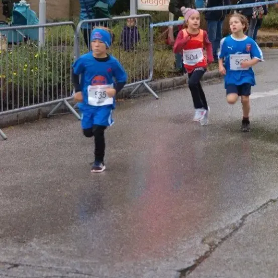 Drei Kinder laufen in einem Rennen mit Nummern auf ihren Trikots. Dahinter hält eine Frau einen Regenschirm und andere schauen zu. Die Straße hat Barrieren und ein Schild.