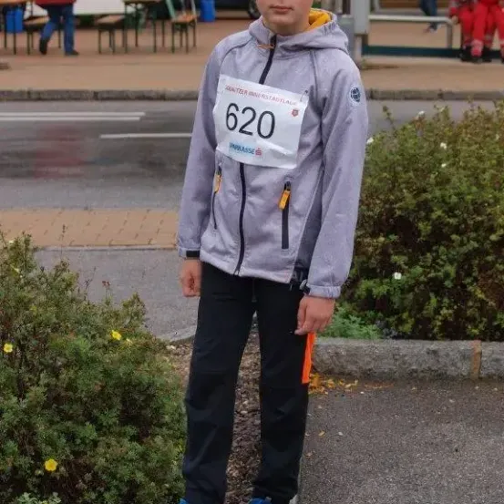 Ein Junge steht auf einem Bürgersteig mit der Nummer 620 auf seiner Jacke, neben einem Busch und einigen Gebäuden.