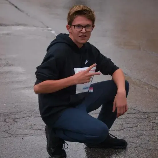 Ein Mann in einer schwarzen Kapuzenjacke und mit Brille hockt auf einer nassen Straße und hält eine weiße Karte.