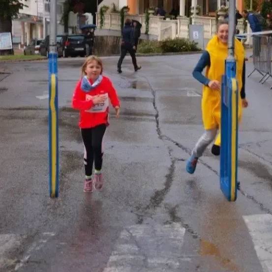 Zwei Läufer, einer in Rot und einer in Gelb, überqueren eine nasse Straße. Hinter ihnen läuft eine Person. Gebäude und geparkte Autos sind sichtbar.