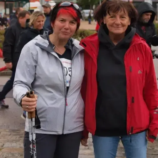 Zwei Frauen stehen zusammen auf einem gepflasterten Bürgersteig, eine hält Skistöcke. Sie lächeln beide. Im Hintergrund gehen mehrere Menschen auf der Straße, und Gebäude und Bäume sind sichtbar.