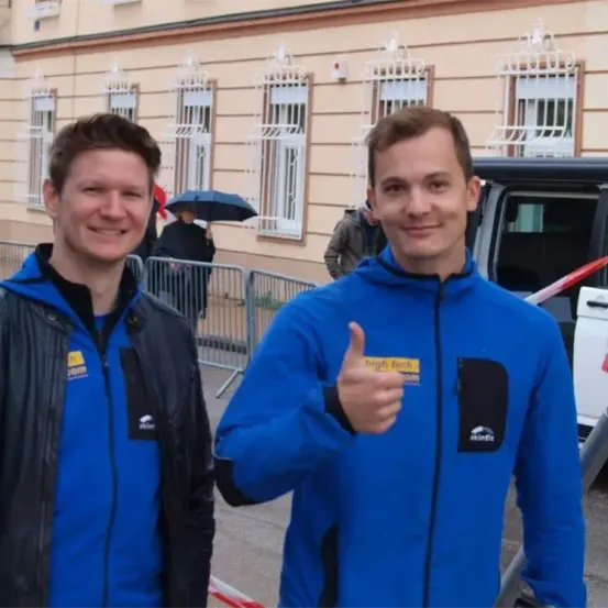 Zwei Männer, einer in einer schwarzen Jacke und der andere in einer blauen Jacke, stehen draußen und geben einen Daumen hoch. Hinter ihnen befindet sich ein Gebäude mit mehreren Fenstern und einem Auto.