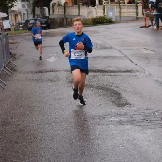Ein Junge in Blau läuft ein Rennen auf einer nassen Straße, während ein anderer Läufer hinter ihm her ist. Zuschauer beobachten von der Seite, und ein Gebäude mit Balkon ist im Hintergrund.