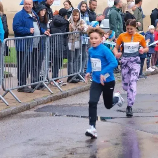 Ein junger Junge läuft bei einem Rennen auf einer nassen Straße, trägt ein blaues Shirt und die Nummer 37. Zuschauer beobachten hinter Barrieren, einige mit Regenschirmen. Ein Mädchen mit der Nummer 735 folgt ihm dicht auf den Fersen.