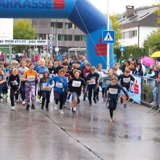 Kinder in Laufausruestung mit nummerierten Shirts laufen bei einem Marathon durch eine nasse Stadtstrasse. Zuschauer und Gebaeude sind im Hintergrund.