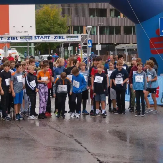 Eine Gruppe von Kindern steht auf einer nassen Straße vor einem Gebäude. Die Kinder tragen nummerierte Trikots und Turnschuhe. Die Straße hat weiße Markierungen und ein Banner mit dem Text 'START-ZIEL'.
