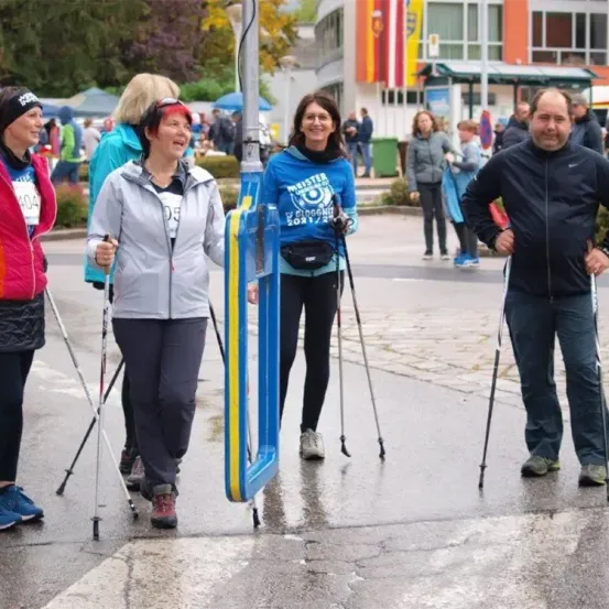 Fünf Personen gehen auf einer nassen Straße und benutzen Trekkingstöcke. Sie tragen Jacken und Turnschuhe. Vor ihnen steht eine blaue Barriere. Dahinter gehen mehrere Personen auf der Straße. Bäume und ein Gebäude sind in der Ferne.