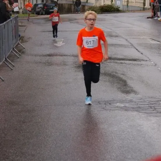 Ein Läufer in einem orangefarbenen Trikot mit der Nummer 617 sprintet nach vorne, gefolgt von einem anderen Läufer in Rot, auf einer nassen Straße mit Zuschauern.