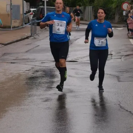 Zwei Läufer nehmen an einem Rennen teil, mit den Nummern 3A und 3B. Sie laufen auf einer nassen Straße, mit Zuschauern. Es gibt auch andere Läufer hinter ihnen.