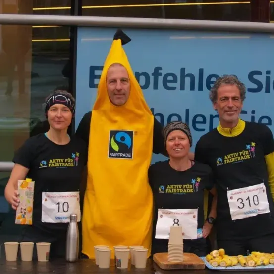 Vier Menschen stehen hinter einem Tisch, zwei Frauen und zwei Männer. Ein Mann trägt eine Bananen-Kostüm. Alle tragen schwarze T-Shirts mit 'Aktiv für Fairtrade'. Sie halten Tassen und Teller mit Essen. Ein blauer Schriftzug hinter ihnen lautet 'Erfohlen Sie helen'.