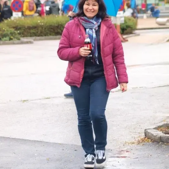 Eine Frau in einer pinken Jacke und blauen Jeans geht im Regen, hält eine Coca-Cola-Flasche und trägt einen Schal.