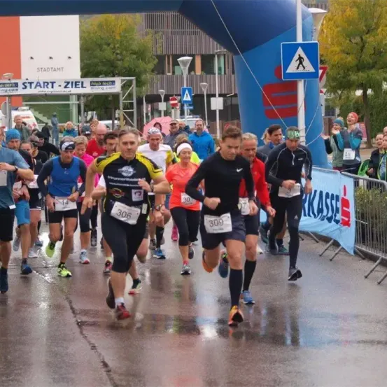 Teilnehmer eines Marathons laufen auf einer nassen Straße und passieren einen blauen Bogen mit Start-Ziel oben. Zuschauer beobachten hinter Metallbarrieren.