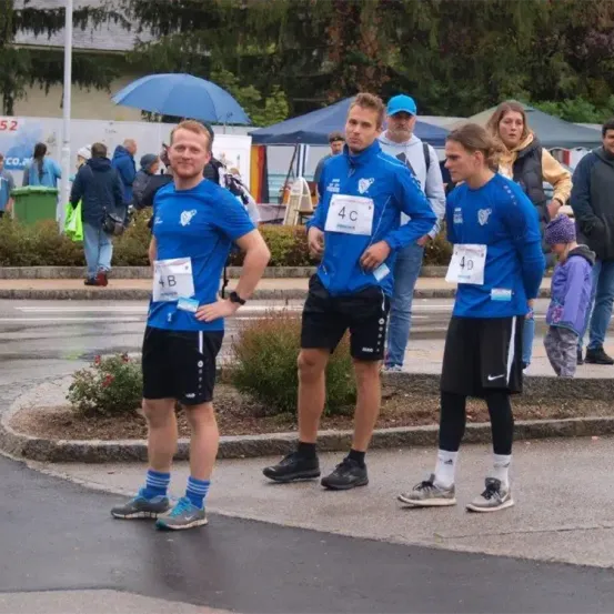 Drei Läufer in blauen Trikots mit Nummern darauf stehen am Straßenrand. Im Hintergrund befinden sich Zelte und Leute, die herumlaufen.