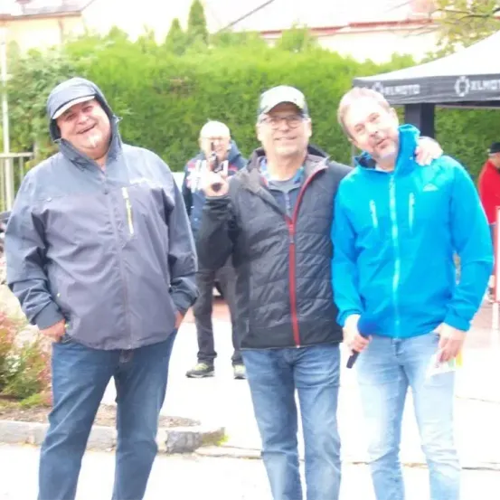 Drei Männer stehen draußen und lächeln. Der Mann links trägt einen grauen Regenmantel, während der Mann in der Mitte eine schwarze Jacke hat. Der Mann rechts ist in einer blauen Jacke. Hinter ihnen ist ein Baldachin mit Logo und Büschen zu sehen.