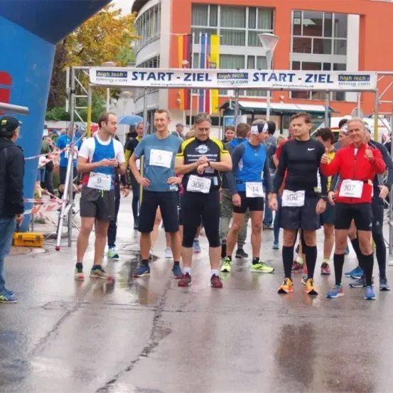 Läufer versammeln sich am Start einer Marathonstrecke, tragen nummerierte Startnummern und Laufschuhe, mit einem Banner oben mit 'START - ZIEL'. Im Hintergrund befinden sich Gebäude und Bäume.