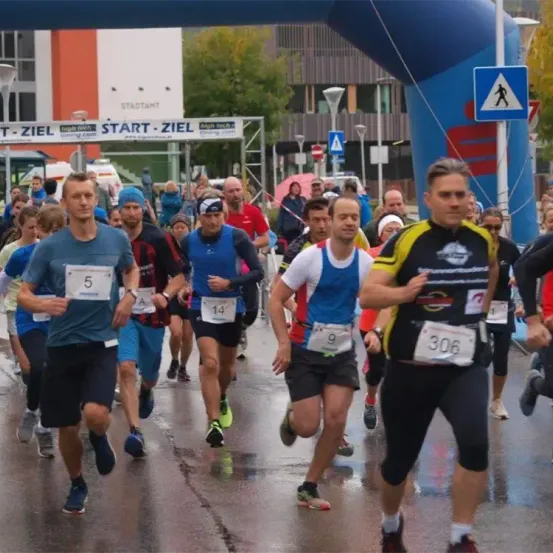 Eine Gruppe von Läufern mit Startnummern nimmt an einem Marathon teil und läuft unter einem Bogen mit der Aufschrift Start-Ziel. Im Hintergrund befinden sich Gebäude und ein blauer Himmel.