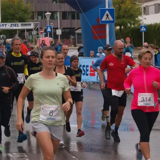 Eine Gruppe von Läufern nimmt an einem Marathon teil. Eine Frau mit der Nummer 6 führt. Dahinter folgt ein Mann in Rot mit der Nummer 23. Zuschauer beobachten von den Seitenlinien aus.