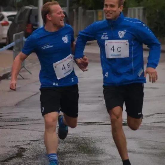 Zwei Männer laufen bei einem Marathon auf einer nassen Straße, beide tragen blaue Trikots und schwarze Shorts. Sie lächeln und zeigen ihre Teilnahme und Kameradschaft.